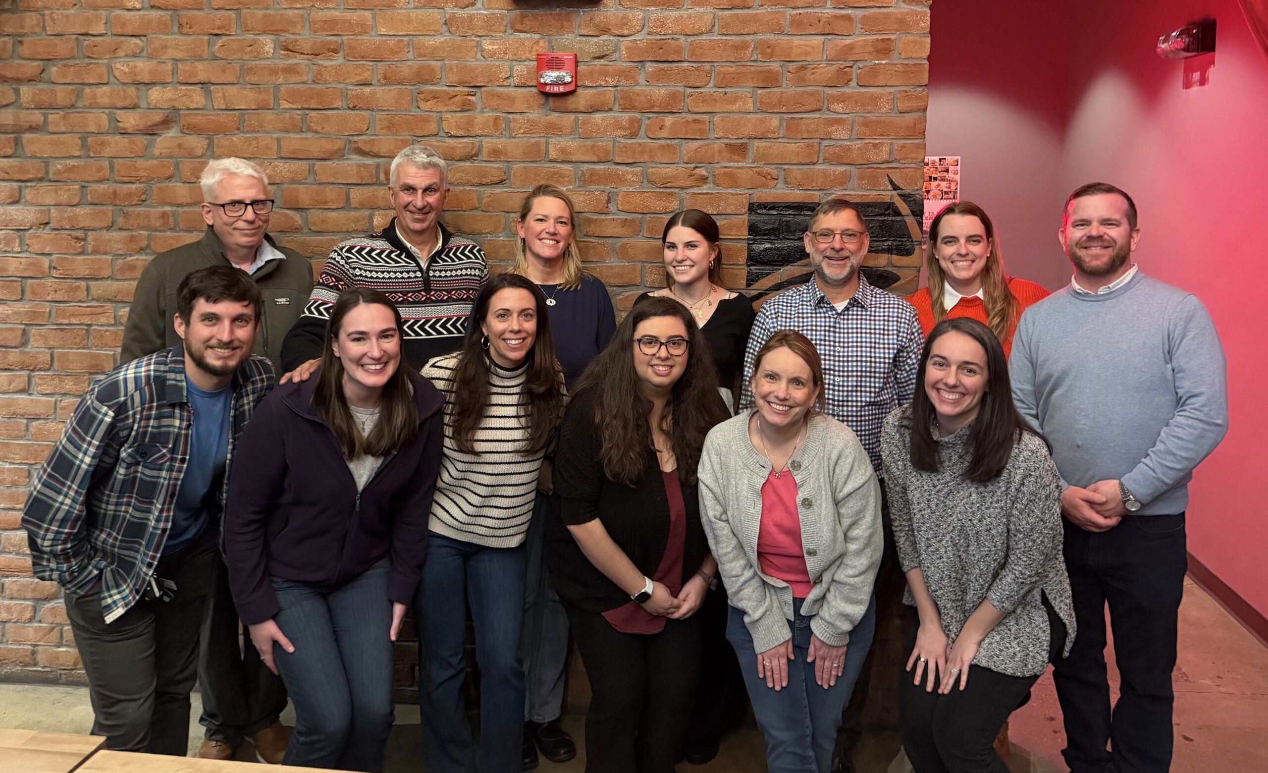 Staff Photo 2025 Triumph Center Group of twelve adults posing together indoors against a brick wall smiling at the camera